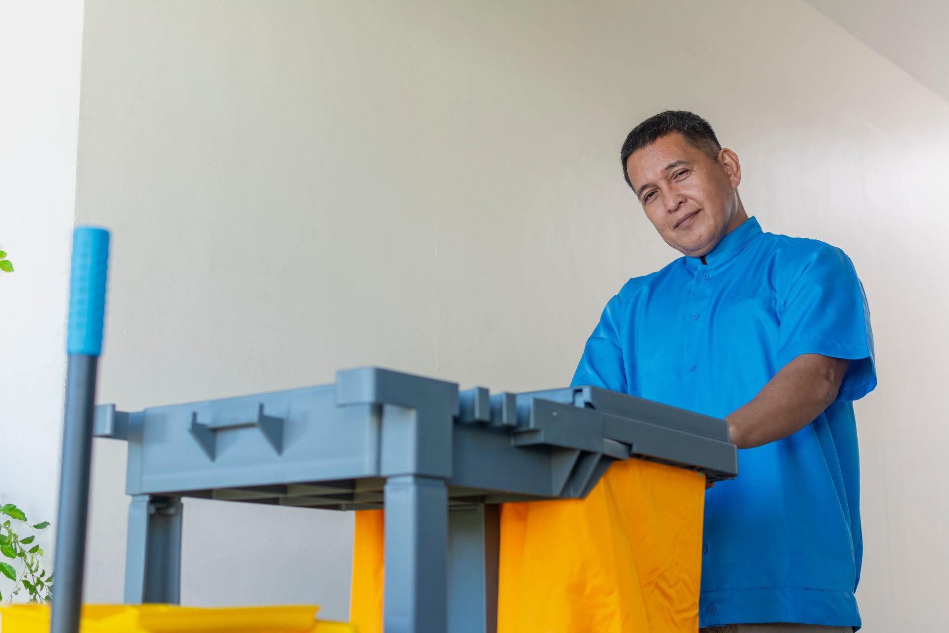A dedicated middle-aged male janitor in a blue uniform pushes a cleaning cart along a bright hallway.